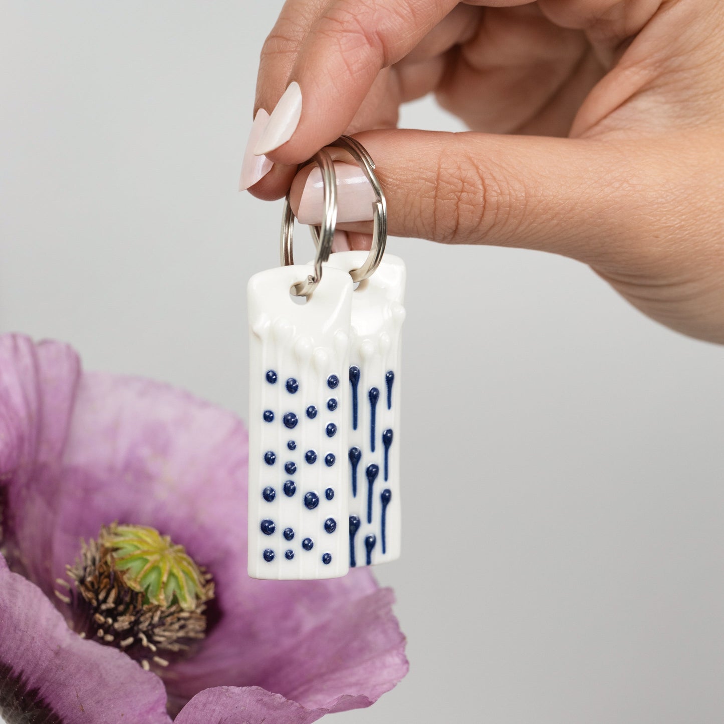 Hand holding handmade porcelain mindful charm keyring with blue slip-trailed tactile decoration beside a pink flower.