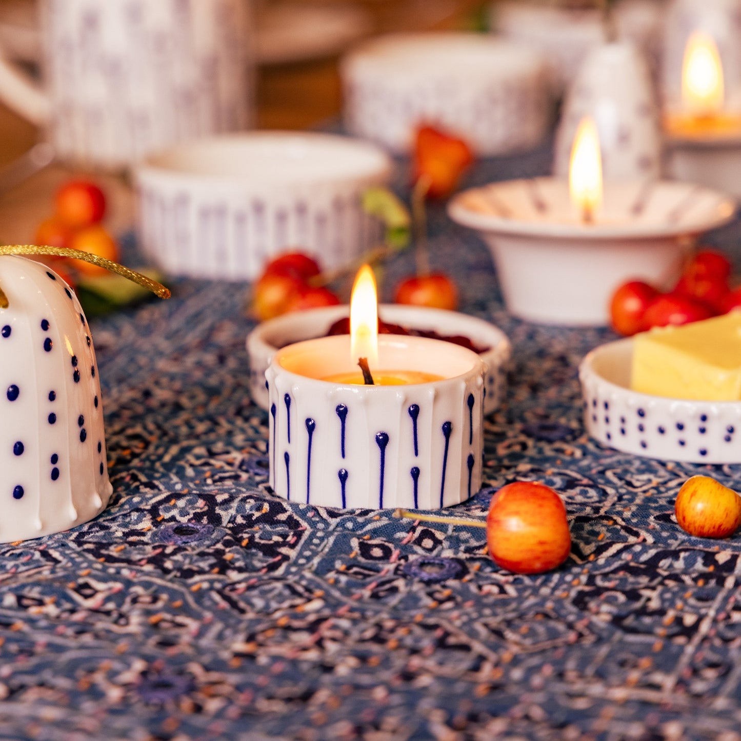 A handmade porcelain tea light holder with blue ink-line decoration, glowing from a beeswax candle on a cozy, patterned table setting.