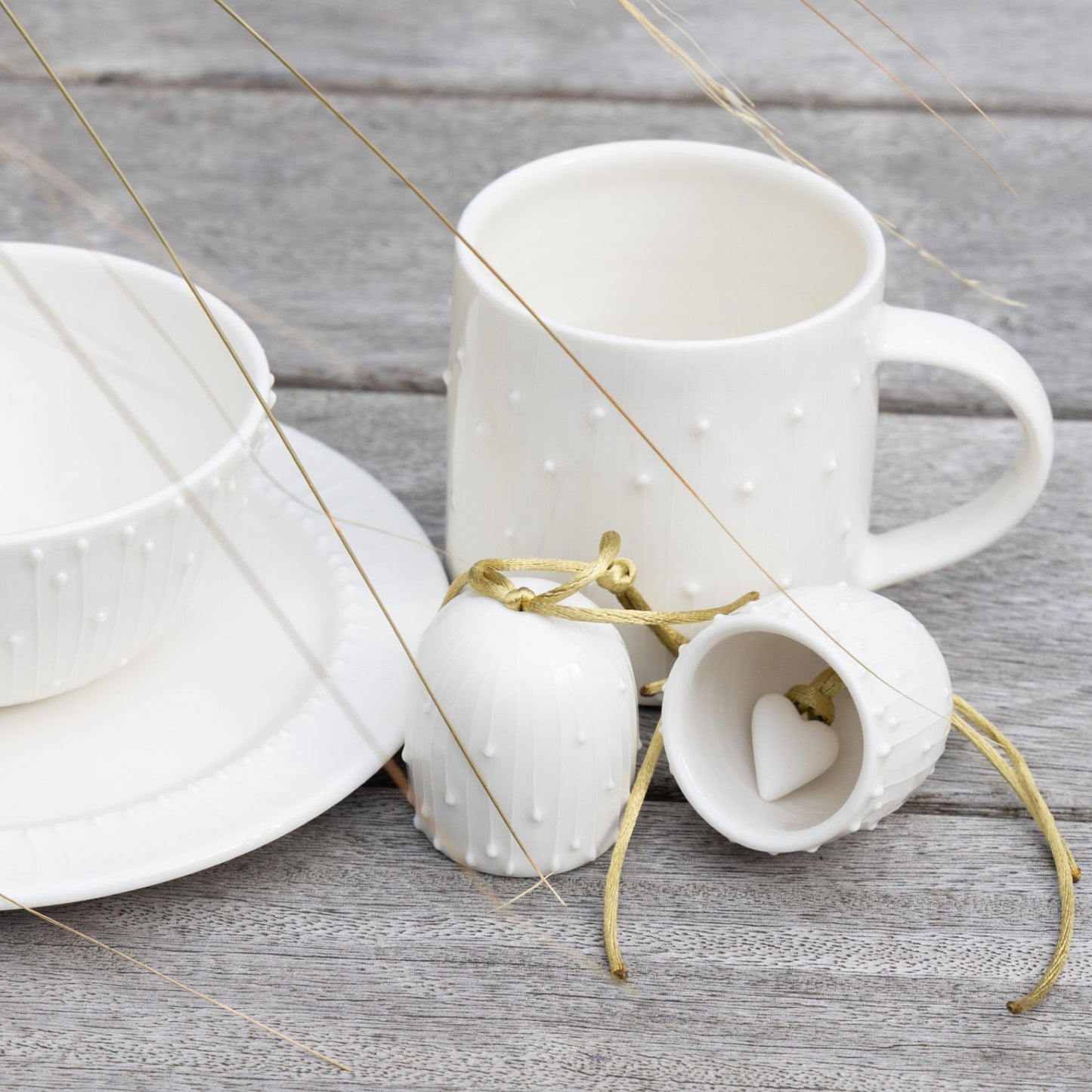 White porcelain mug, bowl, and plate with subtle slip-trailed decoration beside two small porcelain bells