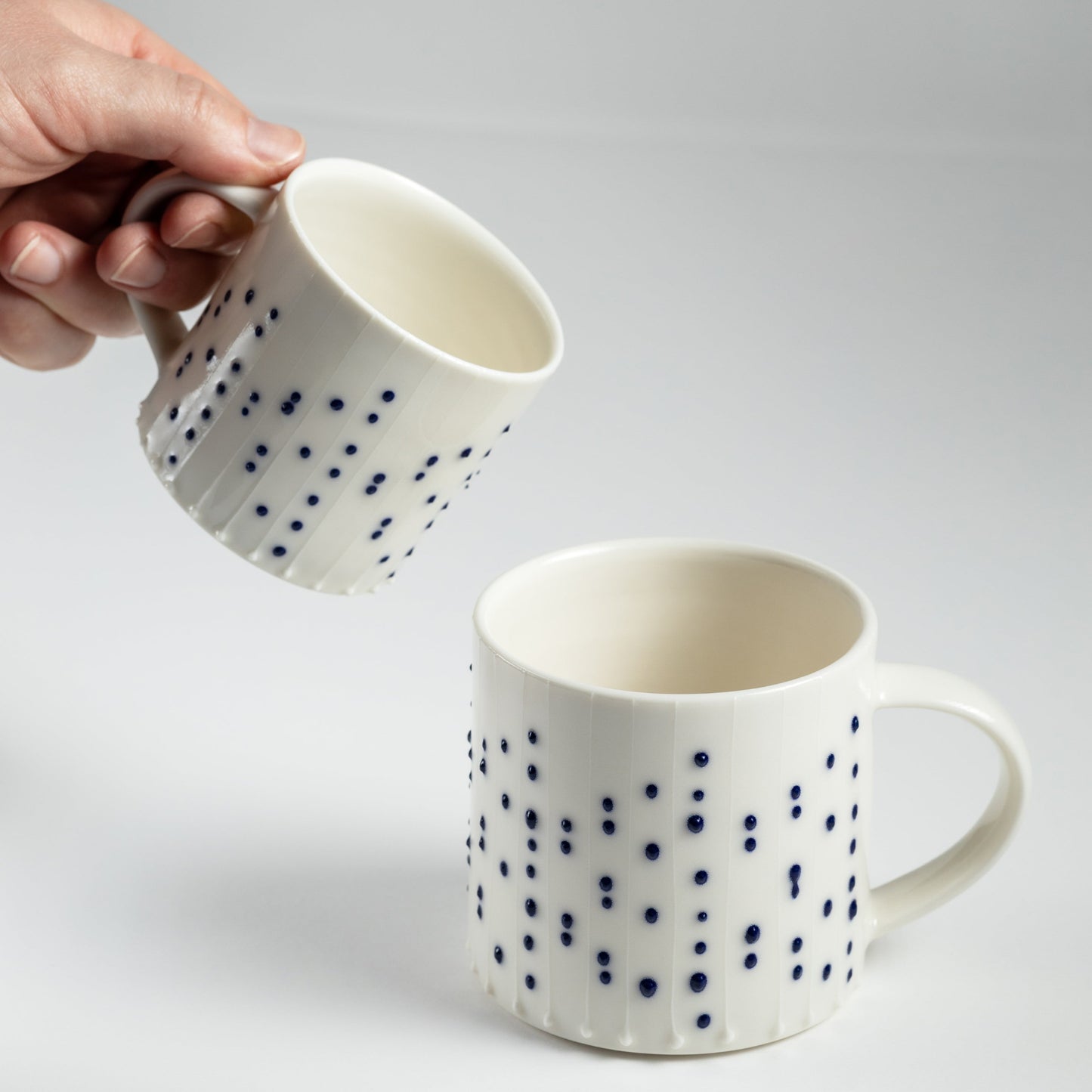 Handmade porcelain small and regular coffee mugs with blue slip-trailed dot decoration, one held in hand
