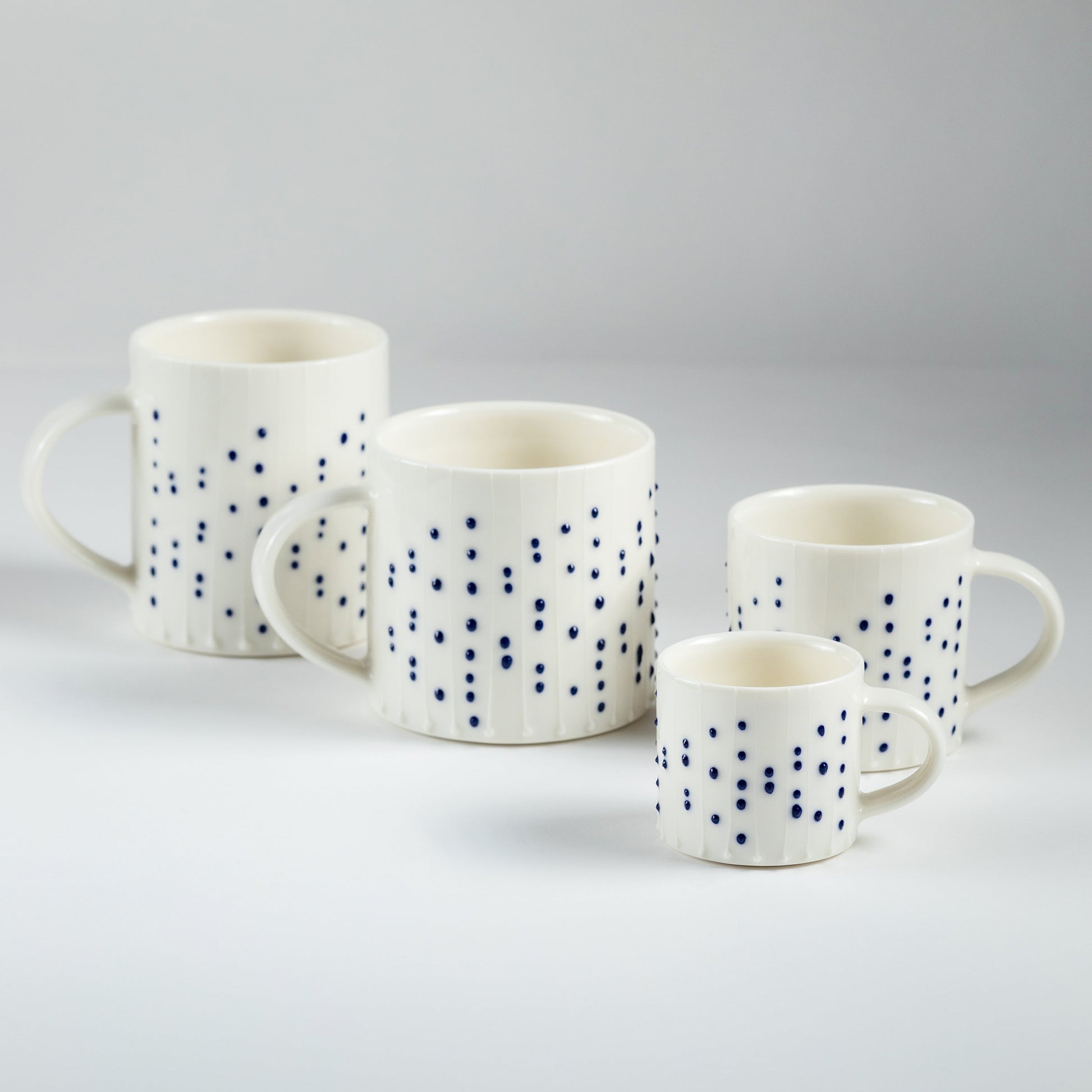 Handmade porcelain espresso cup with blue slip-trailed dot decoration shown beside larger porcelain mugs and cups for size comparison