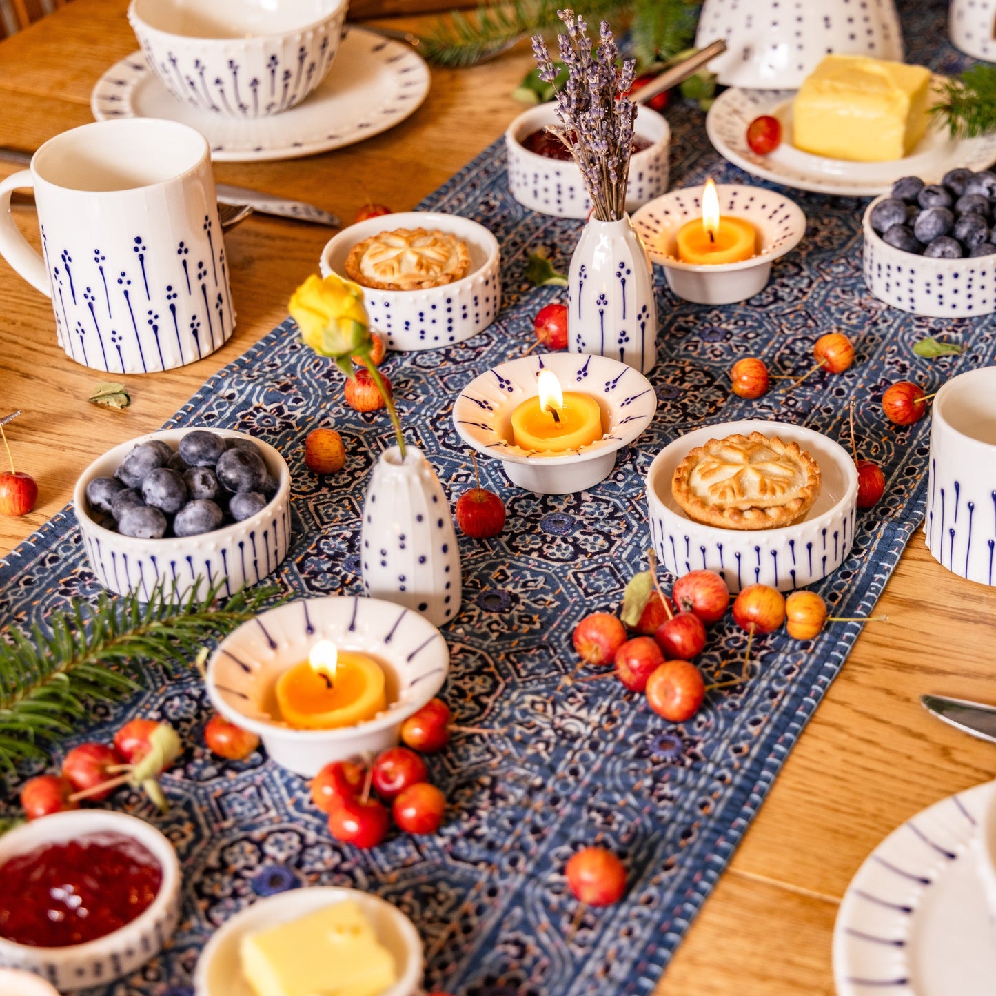 Handmade porcelain tableware with blue slip-trailed decoration styled on a dining table with beeswax tea lights, bowls, mugs, and small bud vases