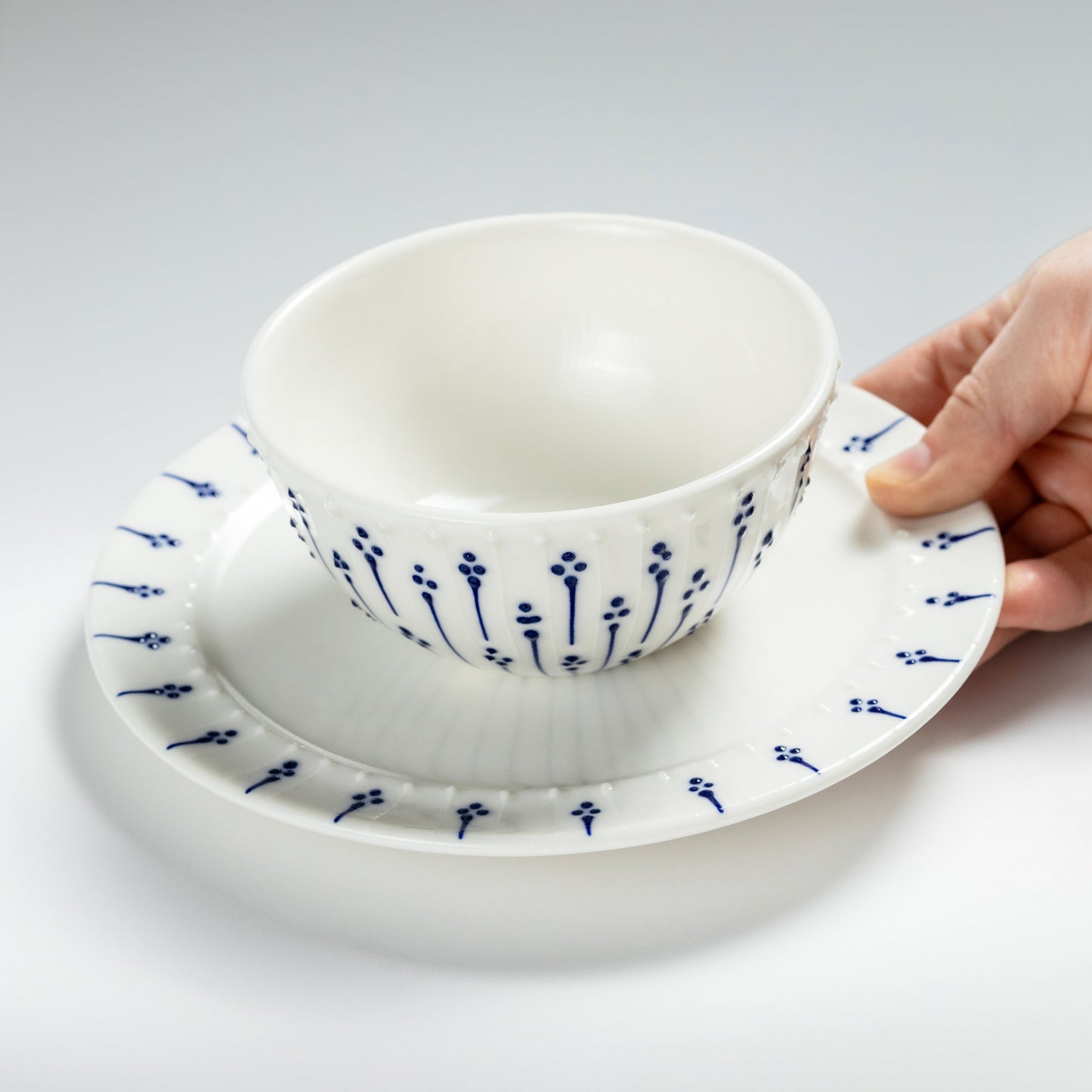 Hand holding handmade porcelain breakfast bowl with blue slip-trailed floral decoration on a matching porcelain side plate