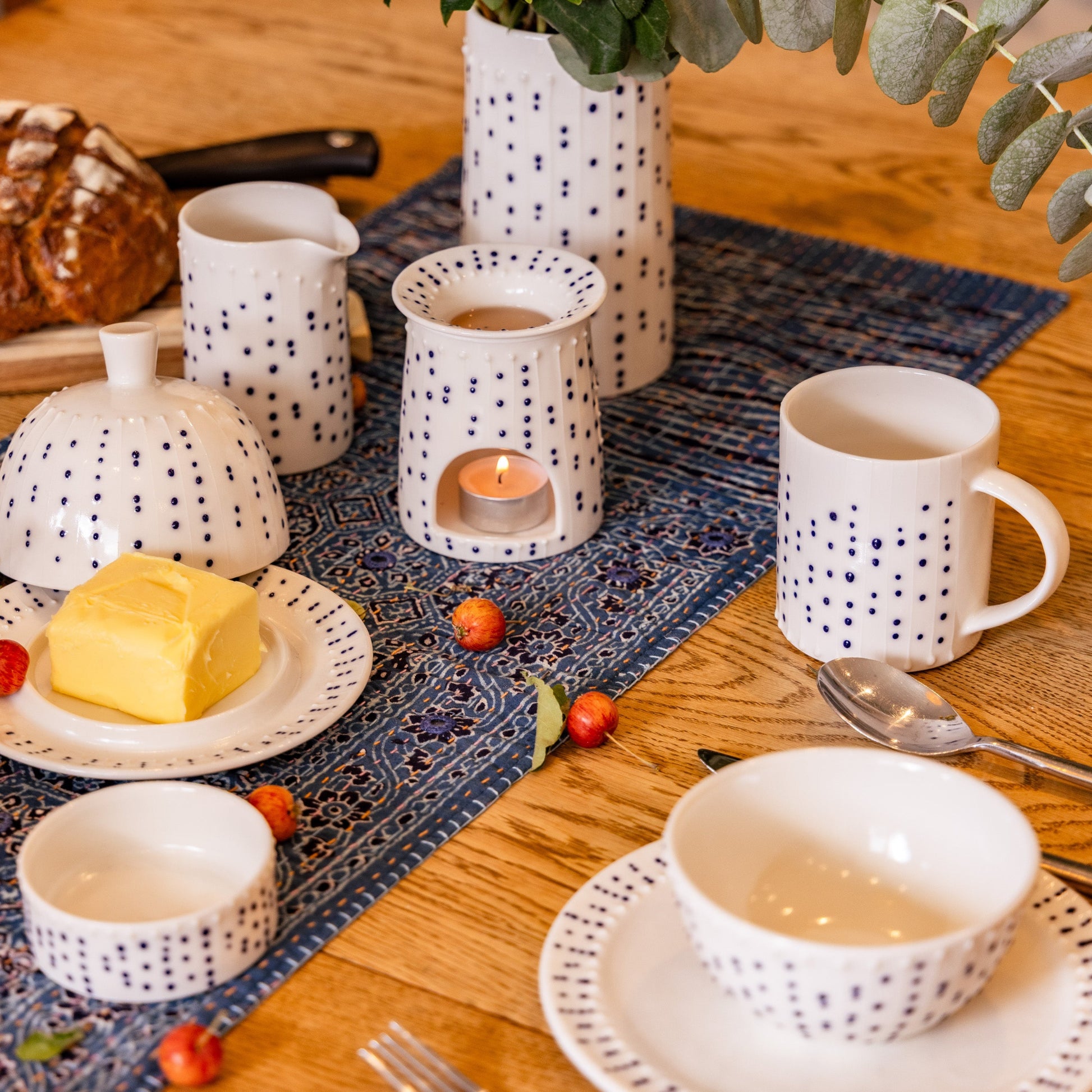 Handmade porcelain tall milk pourer with blue slip-trailed dot decoration on a breakfast table with butter dish, bowls, wax burner, and fresh bread