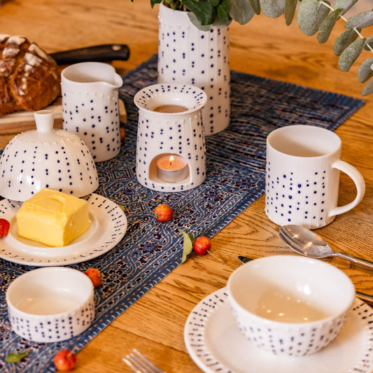Handmade porcelain tall milk pourer with blue slip-trailed dot decoration on a breakfast table with butter dish, bowls, wax burner, and fresh bread