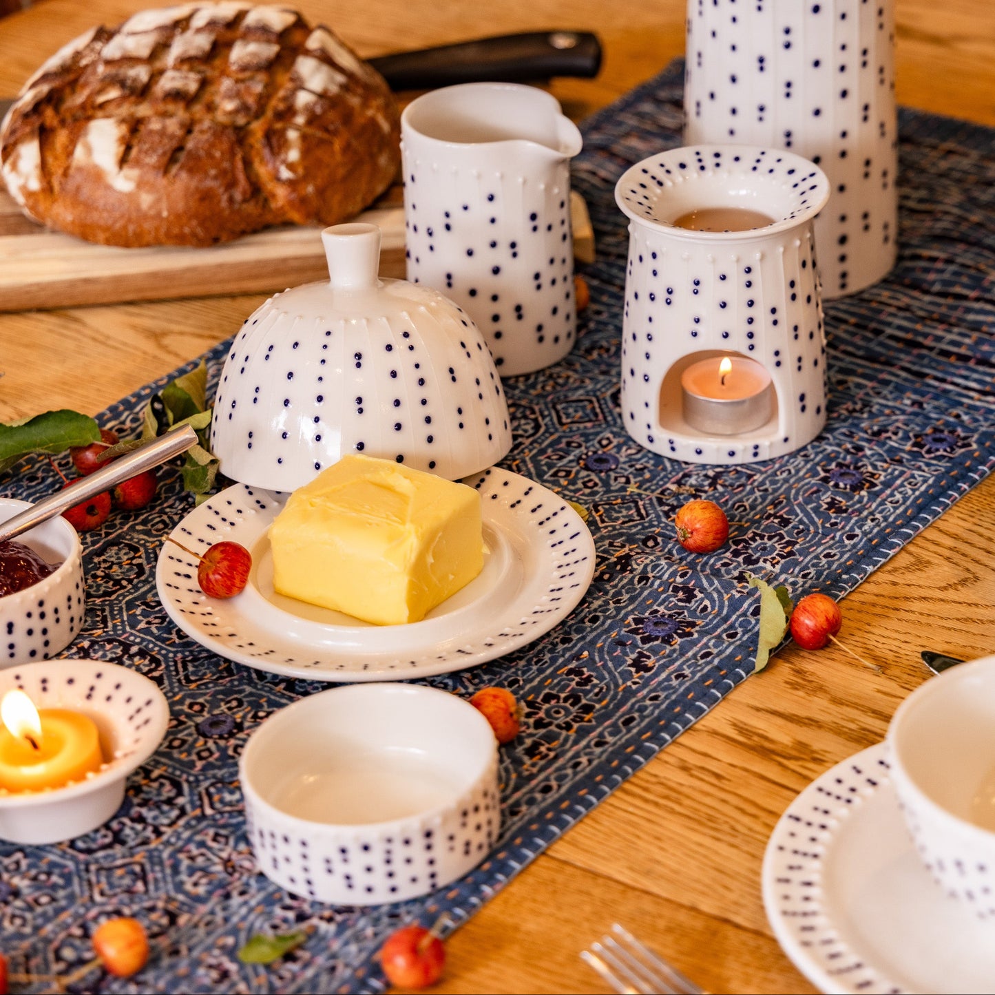 Handmade porcelain butter dish with its domed lid off, revealing a pat of yellow butter, on a wooden table alongside a bread loaf, jam, and a blue pattern table runner