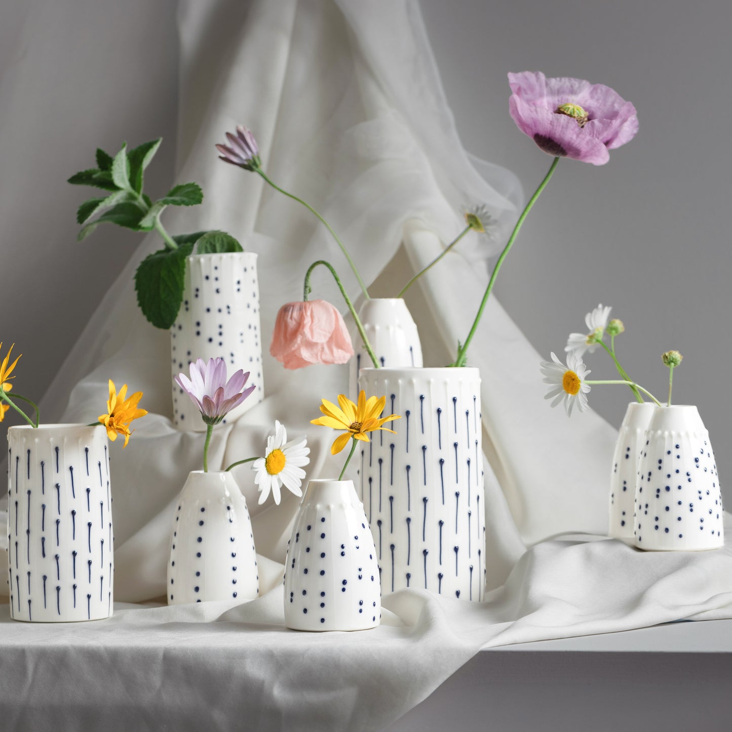 A collection of handmade porcelain flower vases and bud vases in various sizes, decorated with blue ink lines and tactile dots, holding single stems of poppies and daisies.