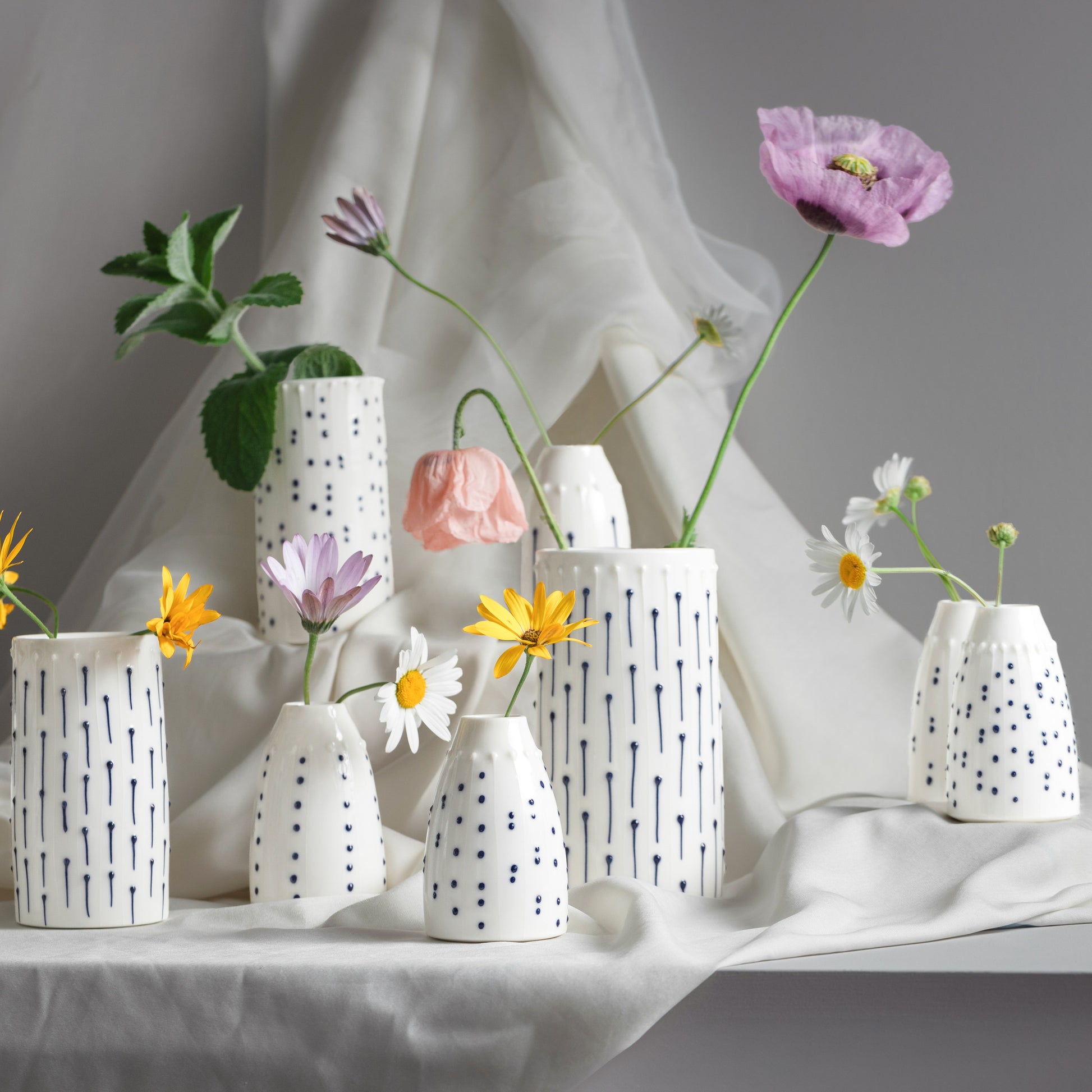 A collection of handmade porcelain flower vases and bud vases in various sizes, decorated with blue ink lines and tactile dots, holding single stems of poppies and daisies.