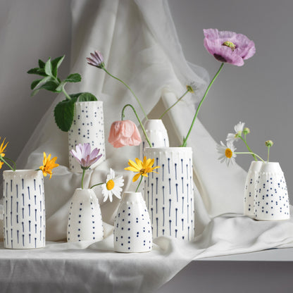 A collection of handmade porcelain flower vases and bud vases in various sizes, decorated with blue ink lines and tactile dots, holding single stems of poppies and daisies.